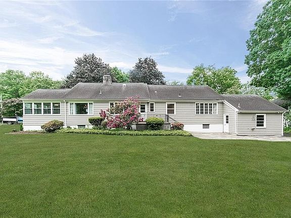 Exterior back picture of 12 Woodside. Please note the small deck off the kitchen with wrought-iron railing and also a flagstone and cement patio.