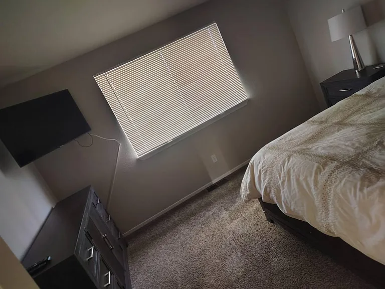 Carpeted bedroom featuring a large window with blinds, a dresser, and space for a full/queen bed and nightstand.