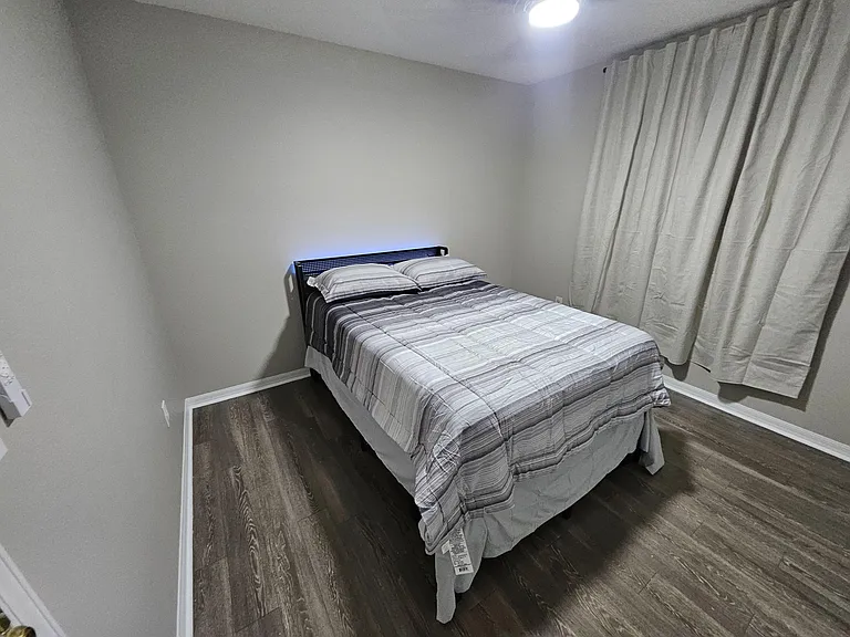 Private bedroom with a full-size bed, wood-look flooring, and a window dressed with full curtains for added privacy.