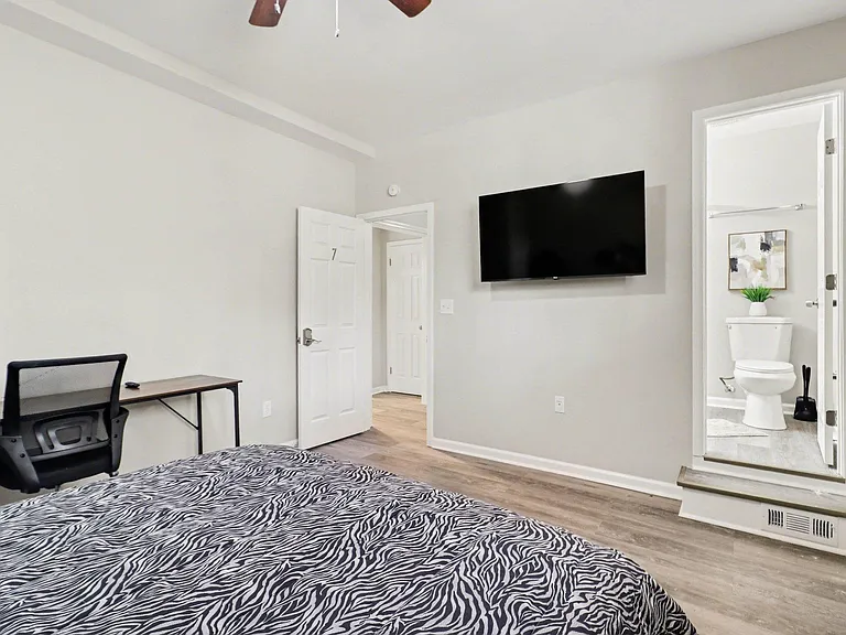Furnished bedroom with mounted TV, work desk, and direct access to a private bath; durable plank flooring throughout.
