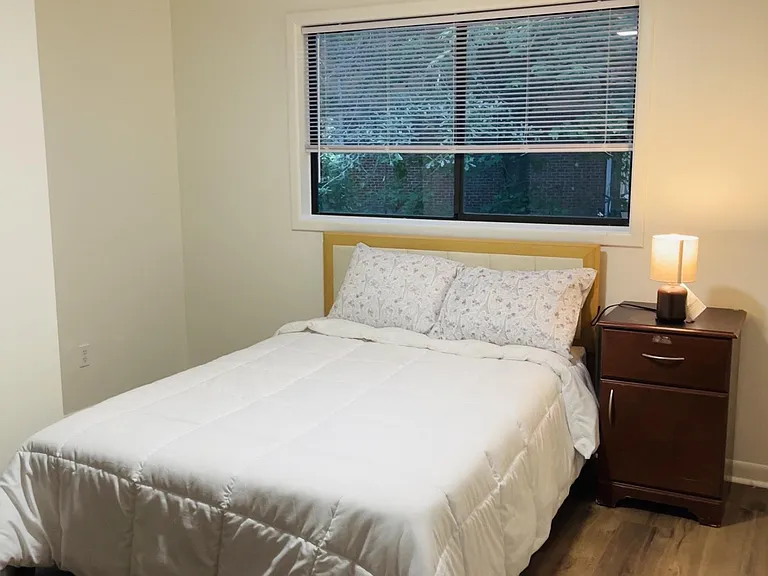 Private bedroom featuring a comfortable bed, nightstand lighting, and a large window with blinds for natural light.