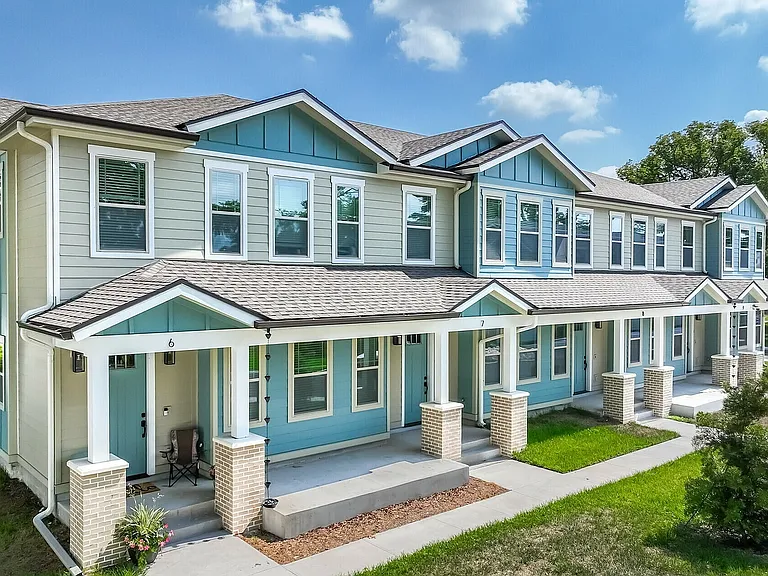Charming townhouse complex with beautiful blue and white exterior, lush landscaping, and a welcoming porch under a bright blue sky.