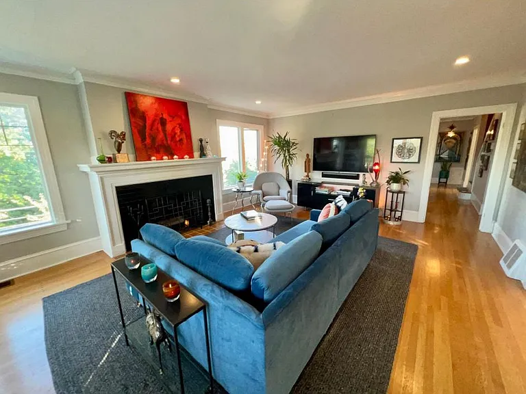 Living room area with white oak flooring and wood burning fireplace. The doorway leads to a hallway and two bedrooms on the main floor and stairs to the second floor.
