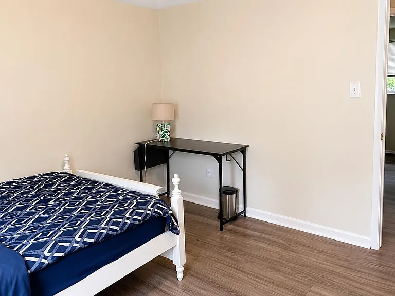 Furnished bedroom with wood-look floors, a work desk with lamp, and easy access to the hallway and nearby window light.