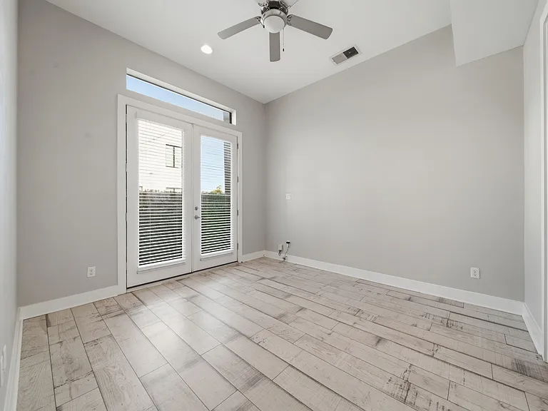 This downstairs bedroom showcases light wood flooring and neutral gray walls for a clean, modern aesthetic. A ceiling fan and expansive glass doors provide excellent natural light and seamless indoor-outdoor access.