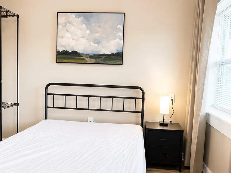 Furnished bedroom with metal bed frame, nightstand and lamp, open shelving, and a window with blinds and curtains.