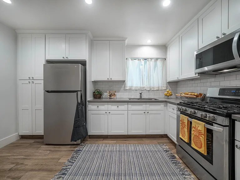 Unit 9 studio gourmet kitchen with stainless steel refrigerator, gas stove & microwave. Recessed ceiling lighting. White cabinetry with gray quartz countertop.