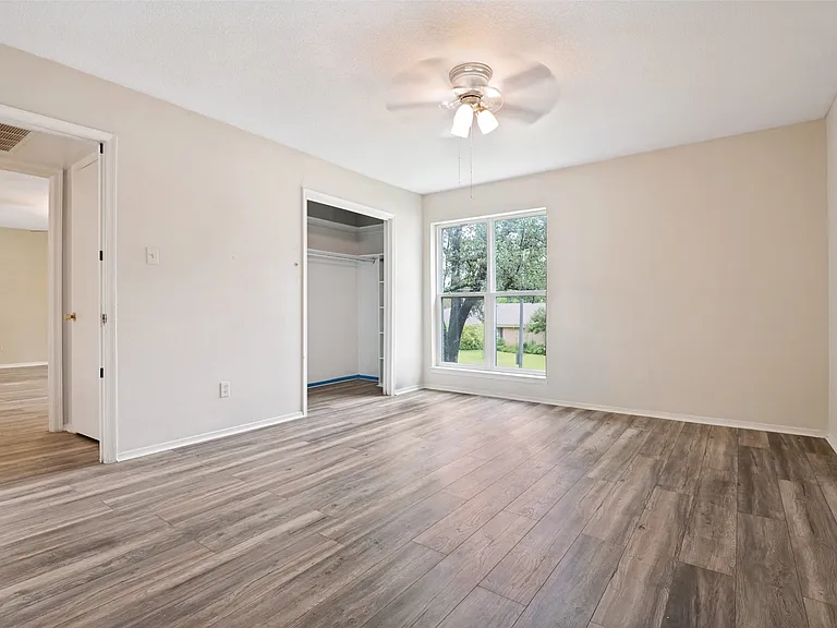 Spacious room featuring wood-finish flooring, a ceiling fan with integrated lighting, and a large window overlooking exterior greenery