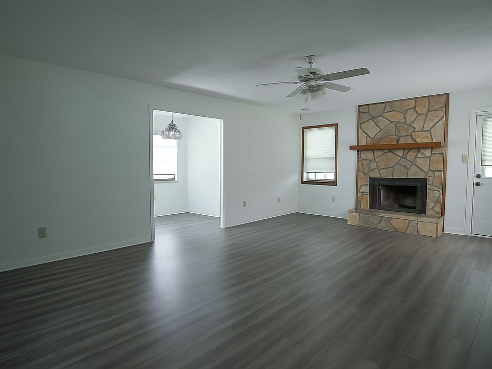 Spacious living area featuring wood-look flooring, a cozy stone fireplace, and open flow to a bright bonus room. Large windows fill the space with natural light, creating a warm and inviting atmosphere ideal for relaxing or entertaining