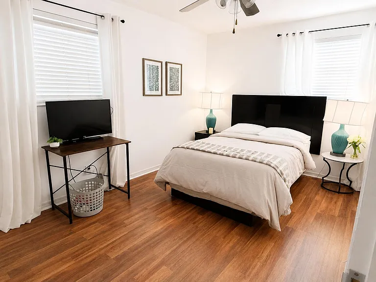 Furnished bedroom with a full bed, nightstands with lamps, TV stand, and two windows with blinds and curtains.