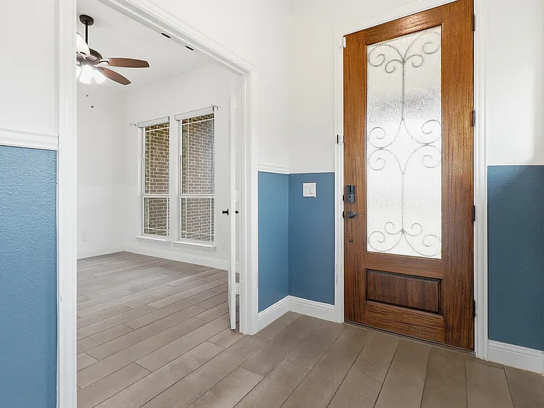 Entryway featuring a wood-panel door with decorative frosted glass, two-tone wall treatment, light wood-finish flooring, and an adjacent room with a ceiling fan and multiple windows