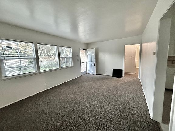 Photo of the living room with the front door and three large windows facing into the beautiful front yard. Living room has new carpet and a heater that you can actually stand near to get warmed up!