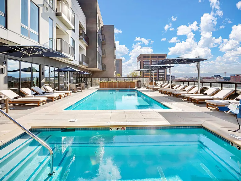 Luxurious rooftop pool area with modern lounge chairs, stylish umbrellas, and stunning cityscape views under a bright blue sky.