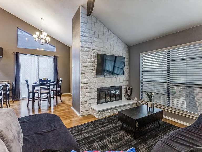 Vaulted ceiling living space featuring a stone-clad fireplace with a white hearth, wood-finish flooring, and a large window with horizontal blinds
