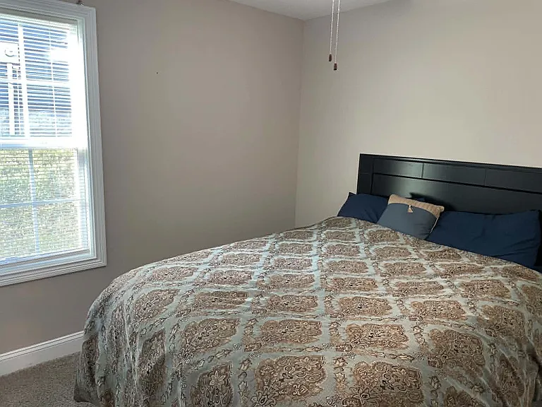 Furnished bedroom with a comfortable bed and a large window with blinds for natural light and privacy.