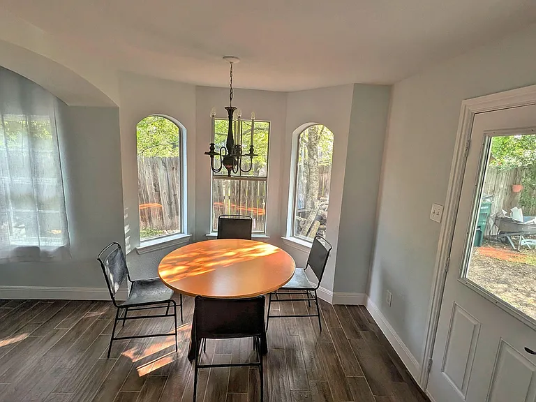 Dining nook features a round table under a chandelier, framed by bay windows with backyard views and durable tile floors.