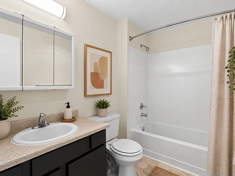 Staged full bathroom with dark vanity cabinetry, granite-style countertop, mirrored medicine cabinet, tub and shower combo, and warm decor accents at Garden Square Apartments in Lakewood, WA. Staged full bathroom with dark vanity cabinetry, granite-style 