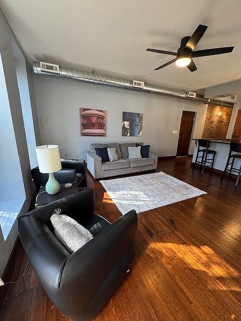 Stylish open-concept living area with a gray sofa, cozy rug, and sleek black chair. The kitchen features warm wood cabinets, stainless steel appliances, and a countertop with barstools. Dark wood floors and exposed ductwork add an industrial-modern vibe.
