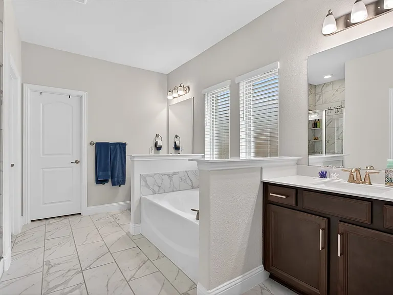 Spacious bathroom featuring a built-in soaking tub with marble-patterned tile surround, dual windows with horizontal blinds, and a double vanity with dark wood cabinetry