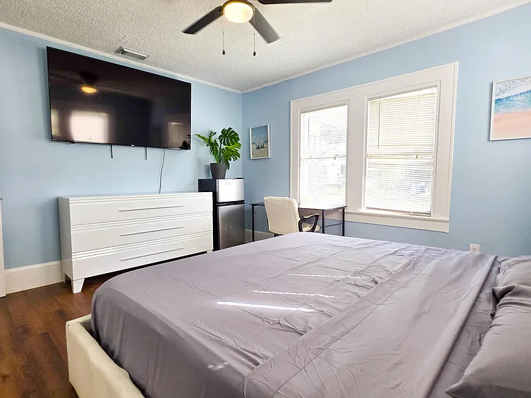 Private bedroom with a full bed, dresser, wall-mounted TV, and a desk by large windows with blinds.