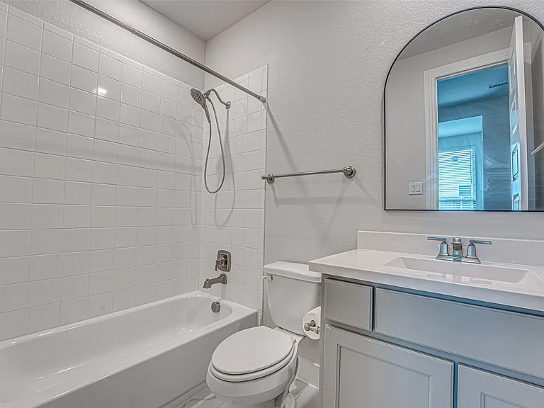 Bathroom featuring a white subway tile shower surround, a white porcelain bathtub, a toilet, a gray vanity with a white countertop and integrated sink, and an arched mirror