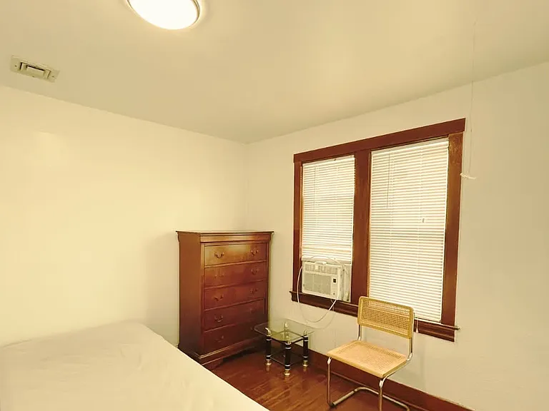 Furnished bedroom with bed, mattress, chair, and dresser for convenient living. Features two large glass windows overlooking an open, unobstructed compound with no nearby buildings, offering a private and relaxed outdoor view with plenty of natural light.